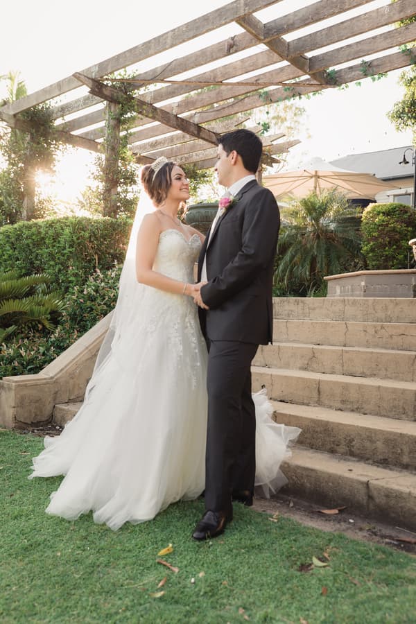 Bride Maryam and groom Pasha stand holding hands near stone steps under a wooden pergola at Hillstone St Lucia — The Quartyard, surrounded by greenery and soft sunlight.