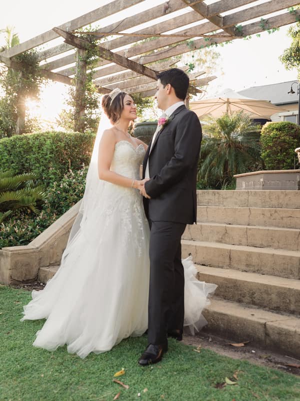 Bride Maryam and groom Pasha stand holding hands near stone steps under a wooden pergola at Hillstone St Lucia — The Quartyard, surrounded by greenery and soft sunlight.