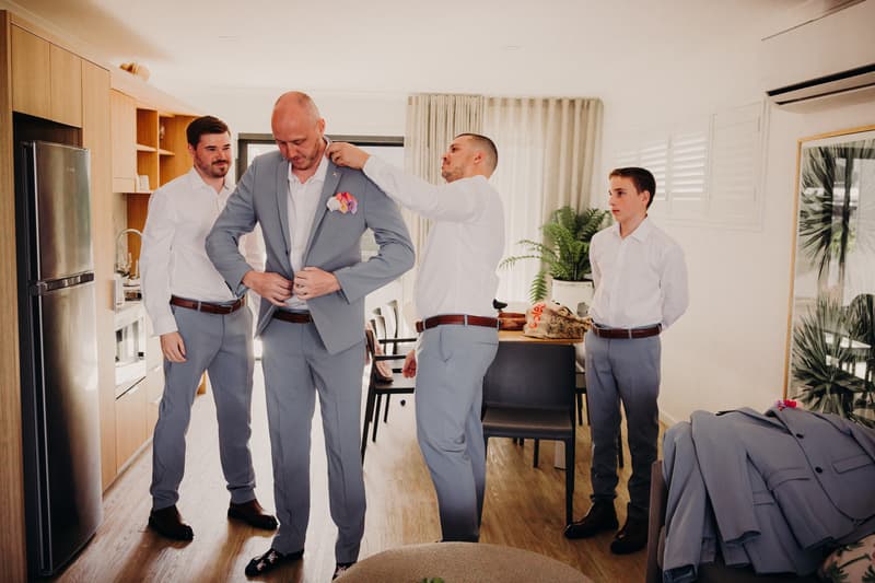 The groom is being assisted with his suit jacket by a groomsman while two other groomsmen stand nearby in a room with kitchen cabinetry and a dining area at Sandstone Point Hotel.