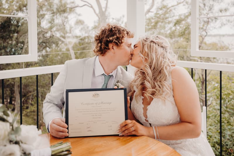 Bride Chloe and groom Brodie kiss while seated at a wooden table holding their framed Certificate of Marriage at Sandstone Point Hotel — Pavilion.