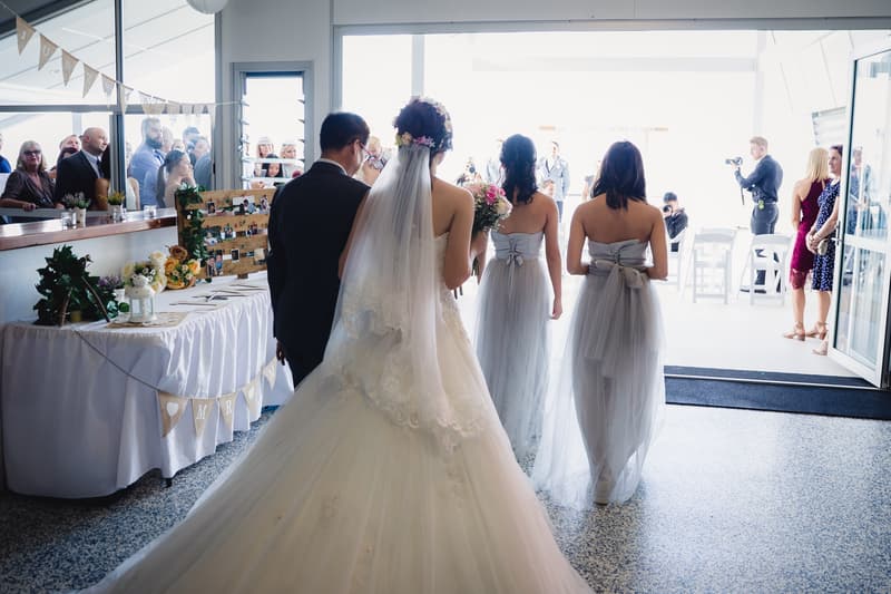 The bride Wing, escorted by an older man, walks down the aisle accompanied by two bridesmaids in grey dresses at Bilinga Beach Weddings — The Terrace, with guests watching and photographers capturing the moment.