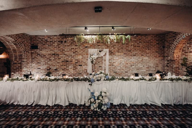 Long reception head table decorated with white and blue floral arrangements, greenery garlands, candles, and draped white fabric at Sandstone Point Hotel — Cellar.