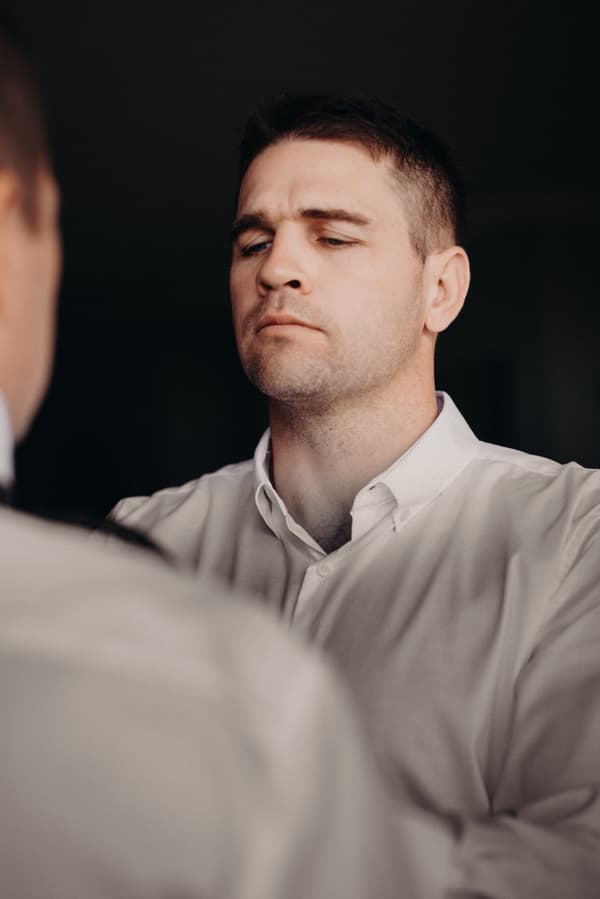 The groom Brent is adjusting his shirt collar while looking into a mirror.