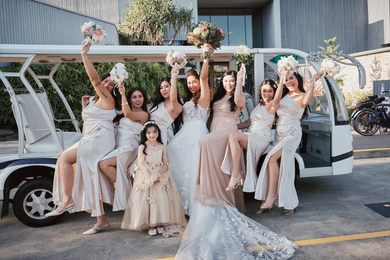 The bride Jheng poses with six bridesmaids and a flower girl outside the Sandstone Point Hotel — The Pavilion, all holding bouquets and smiling in front of a white shuttle vehicle.