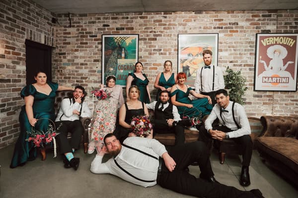Bridal party members pose seated and standing against a brick wall, with bridesmaids in teal dresses and groomsmen in white shirts with black suspenders and bow ties. Engagment party by She Said Yes.