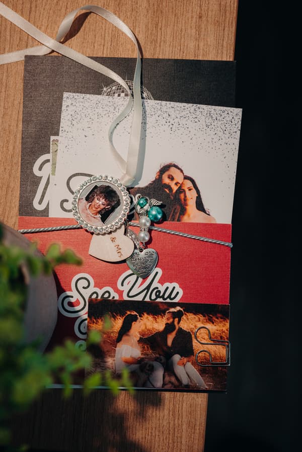 Close-up of wedding stationery and memorabilia on a wooden surface, including photos of the bride and groom, a heart-shaped charm, and a pendant with a portrait.