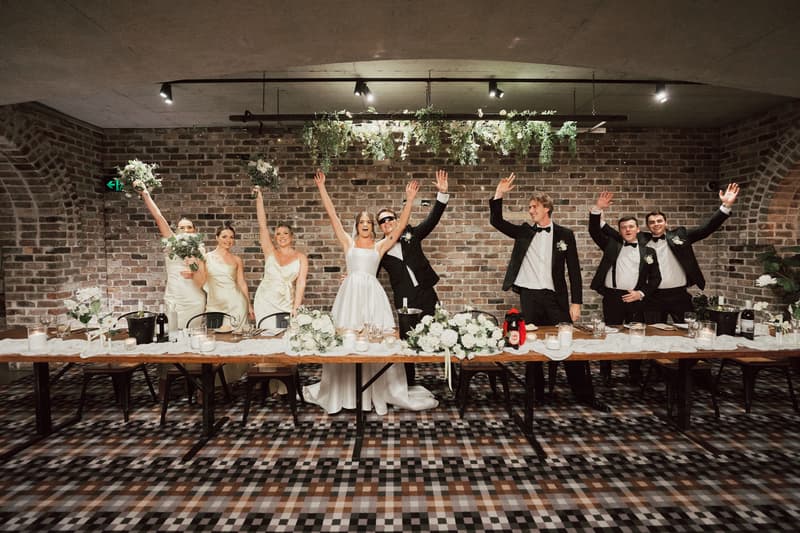 The bride Ashleigh and groom James stand at the center of a long table at Sandstone Point Hotel — The Cellar, surrounded by bridesmaids in cream dresses and groomsmen in black tuxedos, all raising their hands and bouquets in celebration.