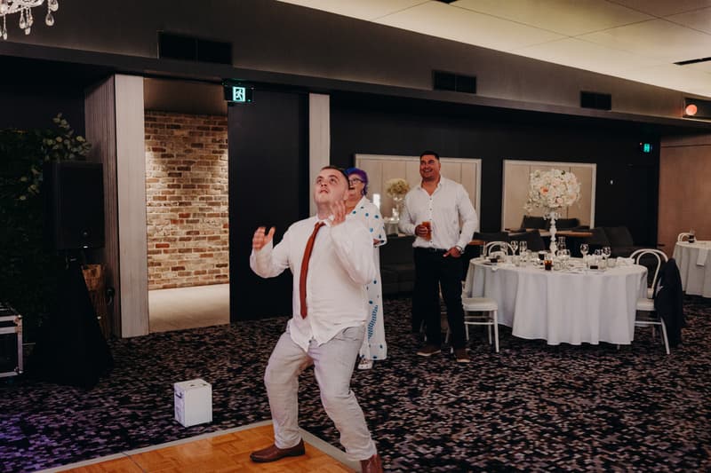 A male guest in a white shirt and red tie is preparing to catch or toss a bouquet or similar object on the dance floor at the Sandstone Point Hotel — Pumicestone Room reception. Two other guests, a woman in a white dress with blue patterns and a man in a white shirt holding a drink, stand nearby watching. A round table with white tablecloth and floral centerpiece is visible in the background.