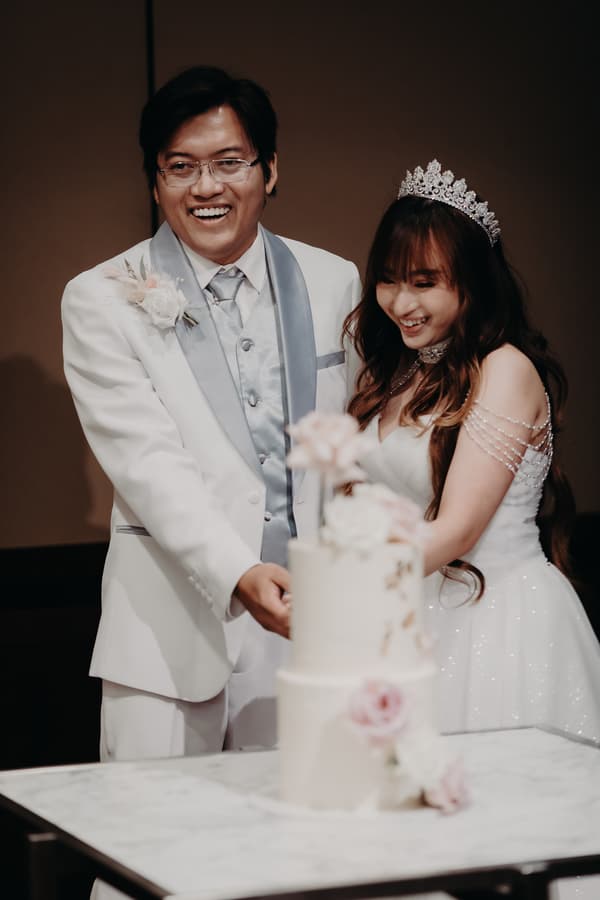 Timothy and Henny cut their wedding cake together at the reception in Royal on the Park. Timothy is wearing a white suit with a silver vest and tie, and Henny is in a white wedding gown with a tiara and pearl shoulder chains.