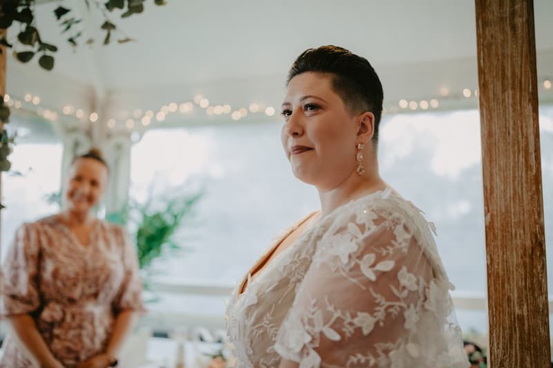 A bride in a white lace dress and earrings stands near a wooden post indoors with string lights and greenery in the background. Another woman in a patterned dress stands blurred in the background.