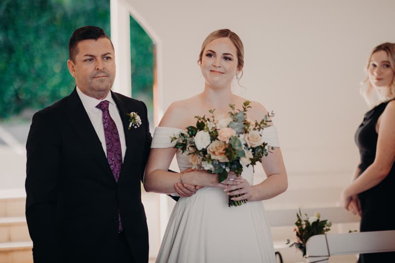 The bride Courtney, holding a bouquet, stands arm-in-arm with an older man, likely her father, inside Tiffany's Maleny — Chapel during the ceremony. A bridesmaid in a black dress is visible in the background.