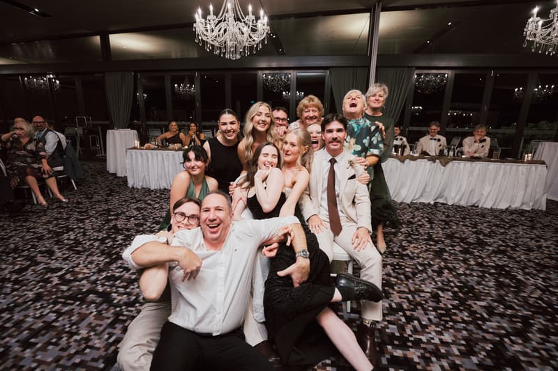 A group of wedding guests pose together on the floor and chairs at the Sandstone Point Hotel — The Pumicestone Room during the reception, with a long head table and chandeliers visible in the background.