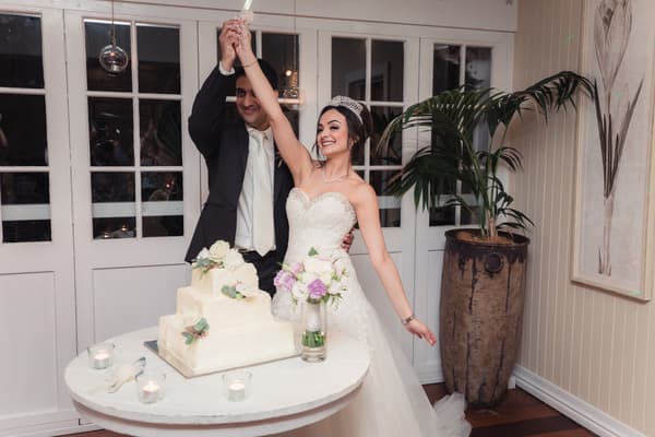 Maryam and Pasha hold a cake knife together above a white wedding cake at Hillstone St Lucia — The Rosewood Room.