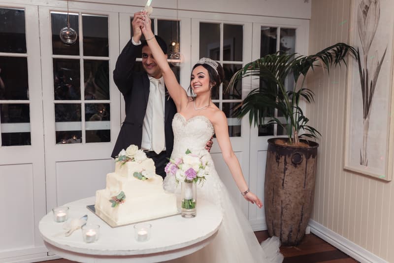 Maryam and Pasha hold a cake knife together above a white wedding cake at Hillstone St Lucia — The Rosewood Room.