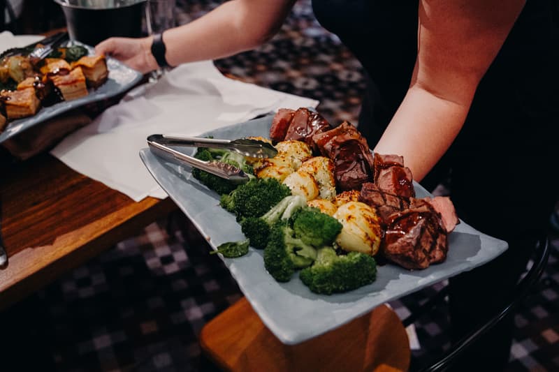 A server holds a rectangular plate with grilled meat, roasted potatoes, and steamed broccoli at the Sandstone Point Hotel — The Cellar during the wedding reception.