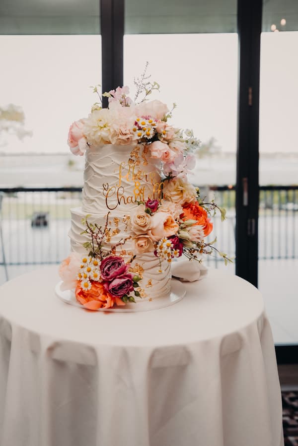 Two-tiered wedding cake decorated with various flowers and gold accents on a round table at Sandstone Point Hotel — Pumicestone Room.