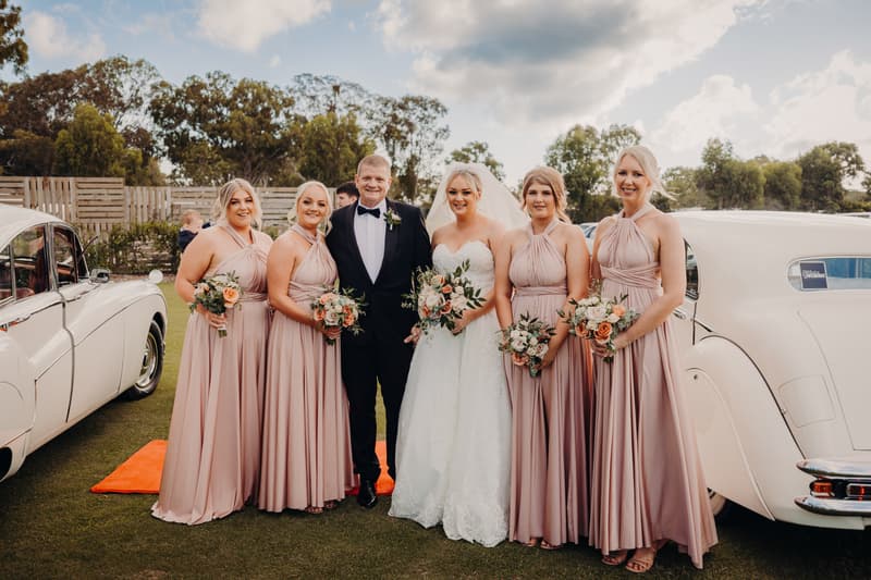 Emily the bride and Dylan the groom pose with four bridesmaids in matching blush pink dresses holding bouquets, standing outdoors between two vintage white cars at Sandstone Point Hotel — Pavilion.