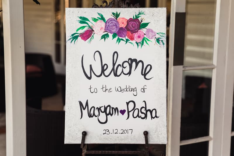 A decorative welcome sign for the wedding of Maryam and Pasha dated 23.12.2017, displayed on an easel with floral artwork at the top.