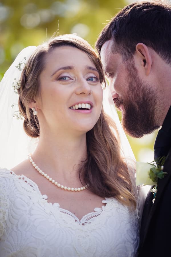 The bride Francesca and the groom Ben share a close moment, with Francesca smiling and wearing a pearl necklace and lace wedding dress, and Ben leaning in close wearing a dark suit with a white rose boutonniere.