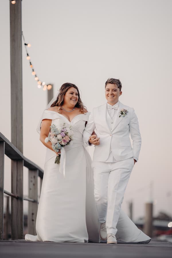 Brooke and Tiffany walk arm in arm outdoors, Brooke holding a bouquet and wearing an off-shoulder wedding gown, Tiffany in a white suit with a boutonniere.