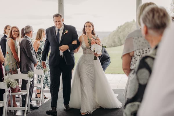 The bride Chelsea walks down the aisle arm-in-arm with her father at Preston Peak Winery Functions Centre, while guests stand and watch.