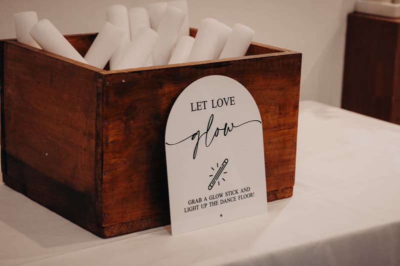 A wooden box filled with white rolled-up glow sticks sits on a table covered with a white cloth at The Tides — The Pandanus Room, accompanied by a sign that reads 'Let Love Glow. Grab a glow stick and light up the dance floor!'