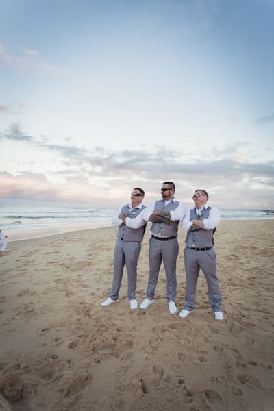 Three groomsmen stand on the beach with arms crossed, wearing grey vests, white shirts, and sunglasses. Destination wedding videographer by She Said Yes.