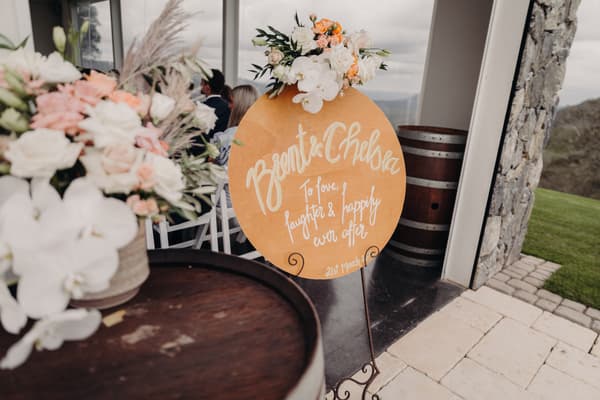 A round wedding sign with floral decoration reads 'Brent & Chelsea To love, laughter & happily ever after 21st March' displayed on a stand near a stone pillar and wooden barrels at Preston Peak Winery Functions Centre. Guests are seated indoors in the background.