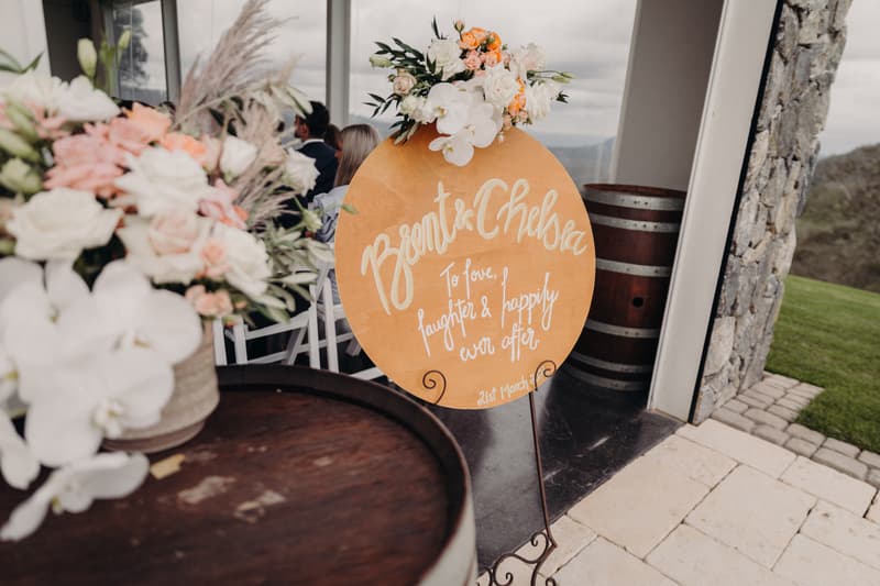 A round wedding sign with floral decoration reads 'Brent & Chelsea To love, laughter & happily ever after 21st March' displayed on a stand near a stone pillar and wooden barrels at Preston Peak Winery Functions Centre. Guests are seated indoors in the background.
