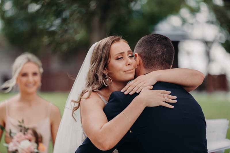 The bride Chelsea embraces the groom Brent during the ceremony at Preston Peak Winery Functions Centre, with a bridesmaid holding a bouquet visible in the background.