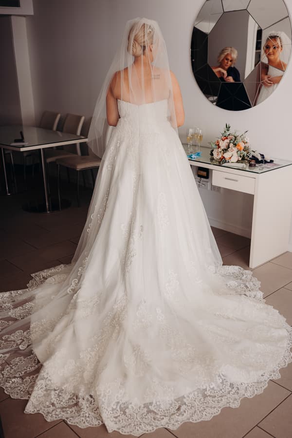 The bride Emily stands with her back to the camera wearing a white lace wedding gown and veil in a room at Sandstone Point Hotel. Her reflection is visible in a round mirror on the wall, showing her face and a woman seated nearby. A bouquet and two glasses of champagne are on a white desk beside her.