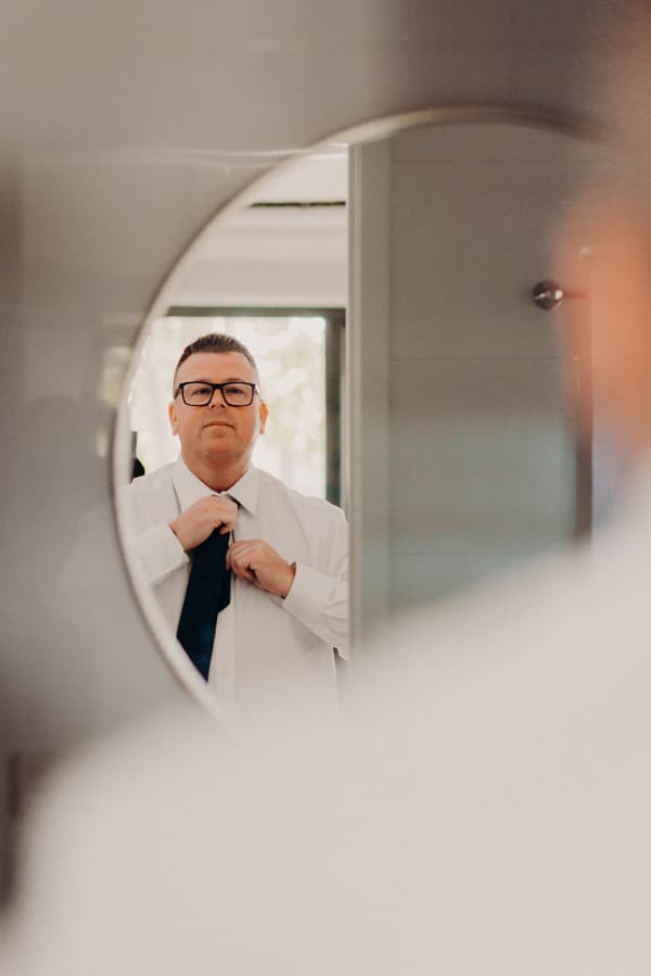 The groom Peter adjusts his tie while looking into a round mirror in a preparation room.