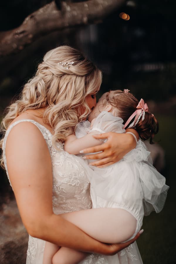 The bride Chloe holds and kisses a young child dressed in white with a pink bow in her hair outdoors at Sandstone Point Hotel.