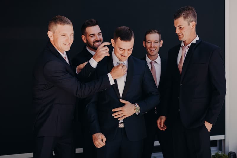 The groom is surrounded by four groomsmen who are adjusting his tie and jacket in preparation at Sandstone Point Hotel.