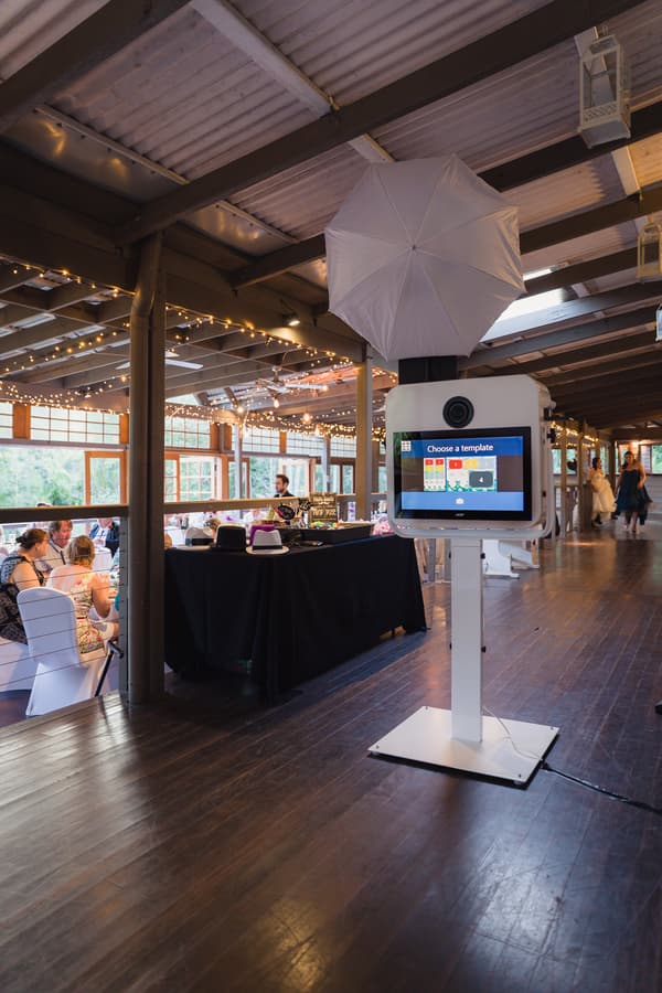 Photo booth setup with a white umbrella light and touchscreen at Kwila Lodge reception, guests seated at tables with fairy lights overhead, and a bride and another guest walking in the background.
