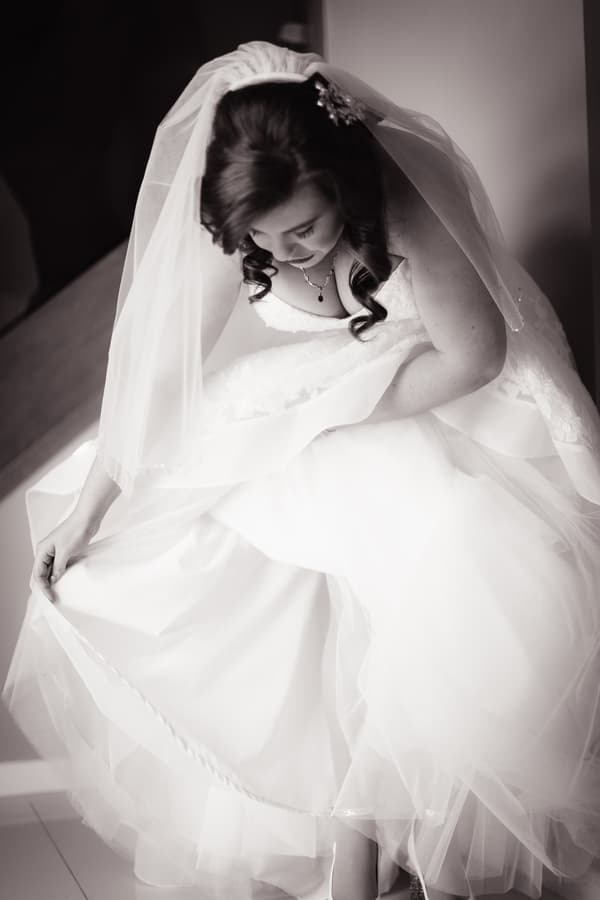 The bride Holly adjusts the hem of her wedding dress while seated, wearing a veil and necklace.