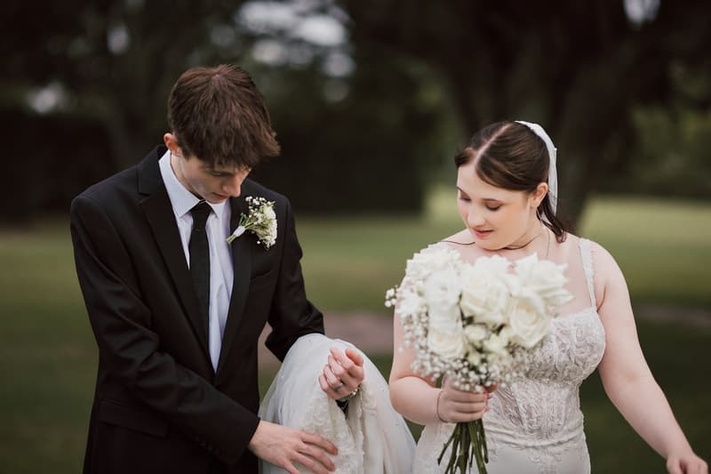 Terion and Tyler stand outdoors at Sandstone Point Hotelallo, with Tyler holding part of Terion's wedding dress while Terion holds a bouquet of white flowers.