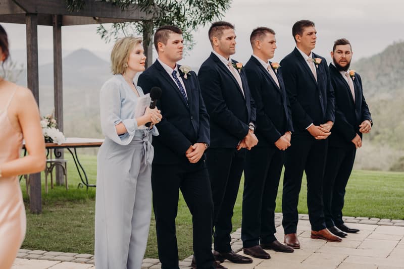 A woman holding a microphone stands next to five groomsmen in dark suits with boutonnières at Preston Peak Winery Functions Centre during the wedding ceremony stage.