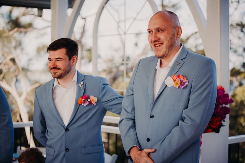 The groom Arran and a groomsman stand inside the Pavilion at Sandstone Point Hotel during the ceremony, both wearing light blue suits with colorful boutonnieres.