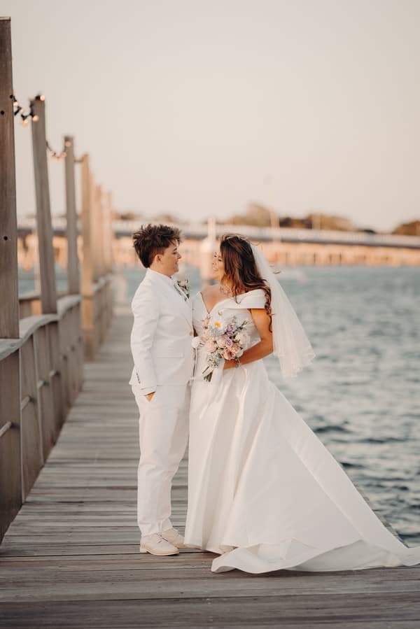 Brooke and Tiffany stand facing each other on a wooden pier by the water, with Brooke wearing a white wedding gown and veil holding a bouquet, and Tiffany dressed in a white suit.