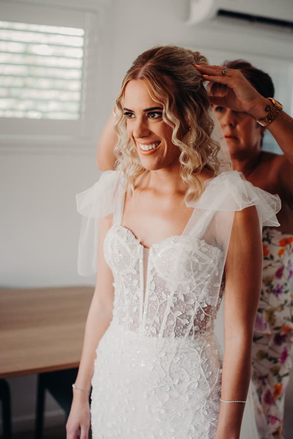 The bride Krystal is smiling while another woman, possibly her mother, adjusts her veil indoors.