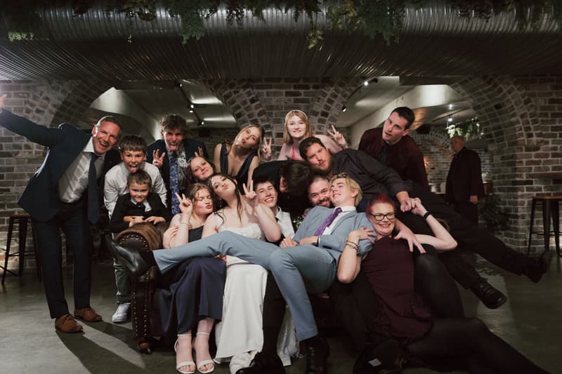 Terion and Tyler pose with a large group of guests on a couch at the reception stage in Sandstone Point Hotel — The Cellar, with brick archways and industrial ceiling visible.