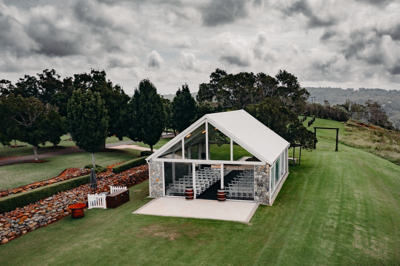 Empty ceremony tent with rows of white chairs and a black aisle runner at Preston Peak Winery Functions Centre, surrounded by green lawn and trees under a cloudy sky.