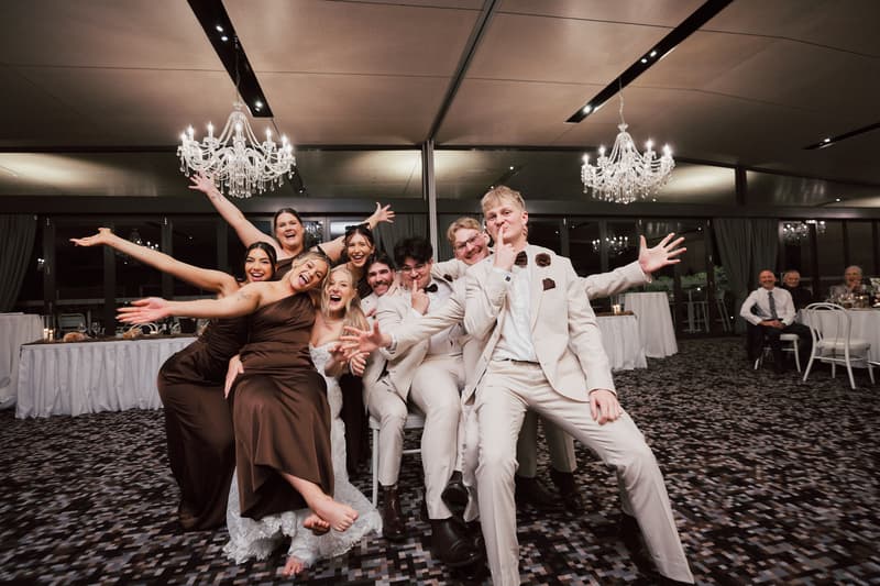 Morgan the bride, Joel the groom, and their bridal party pose playfully on the reception stage at Sandstone Point Hotel — The Pumicestone Room.