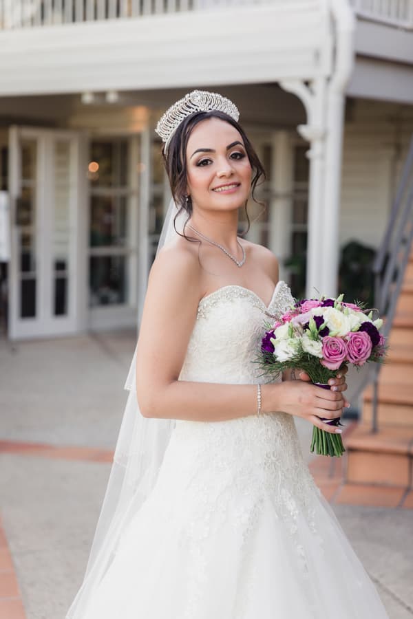 The bride Maryam in a strapless white wedding gown and tiara holds a bouquet of pink, white, and purple flowers outside Hillstone St Lucia, near a building entrance with glass doors and stairs.