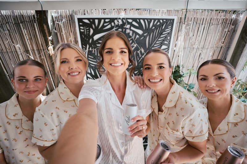 The bride Ashleigh in a white pajama set takes a selfie with four bridesmaids in matching floral pajama sets at Sandstone Point Hotel.