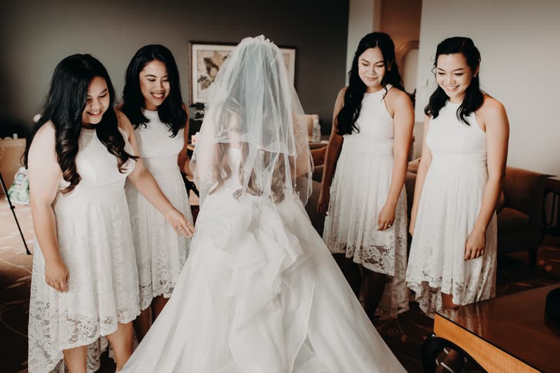 The bride stands with her back to the camera wearing a wedding gown and veil, surrounded by four bridesmaids in matching white lace dresses inside a room at Royal on the Park.