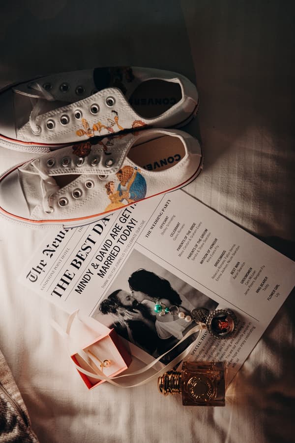White Converse sneakers with Beauty and the Beast design placed on a bed next to a wedding program for Mindy and David at Sandstone Point Hotel, a ring in a pink box, a pearl bracelet, and a perfume bottle.