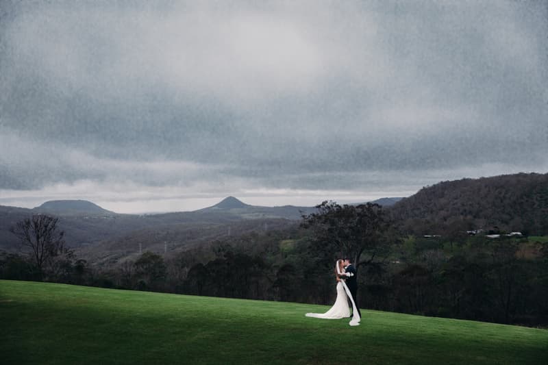 Chelsea and Brent stand together on a grassy hill at Preston Peak Winery Functions Centre with a mountainous landscape and cloudy sky in the background.