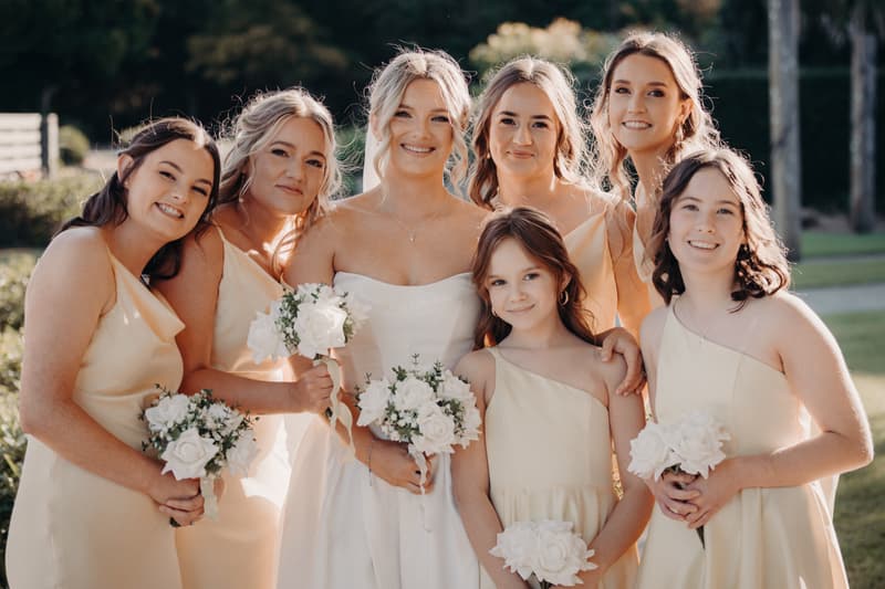 The bride Libby poses with six bridesmaids holding white floral bouquets outdoors at The Tides — The Water's Edge.